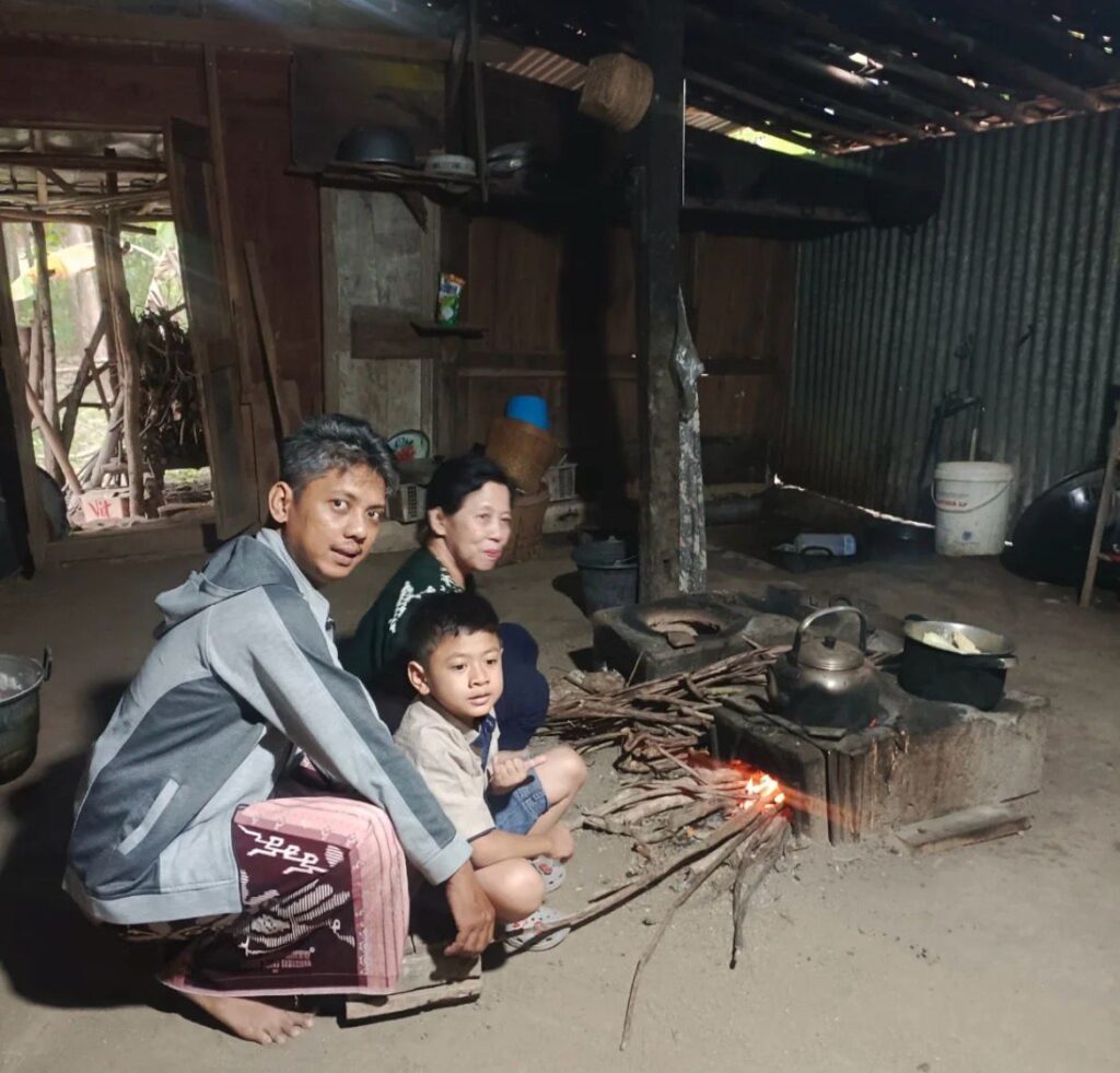 Family sitting by a small open fire stove inside a rustic home with a kettle on the fire and a boy a man and a woman present